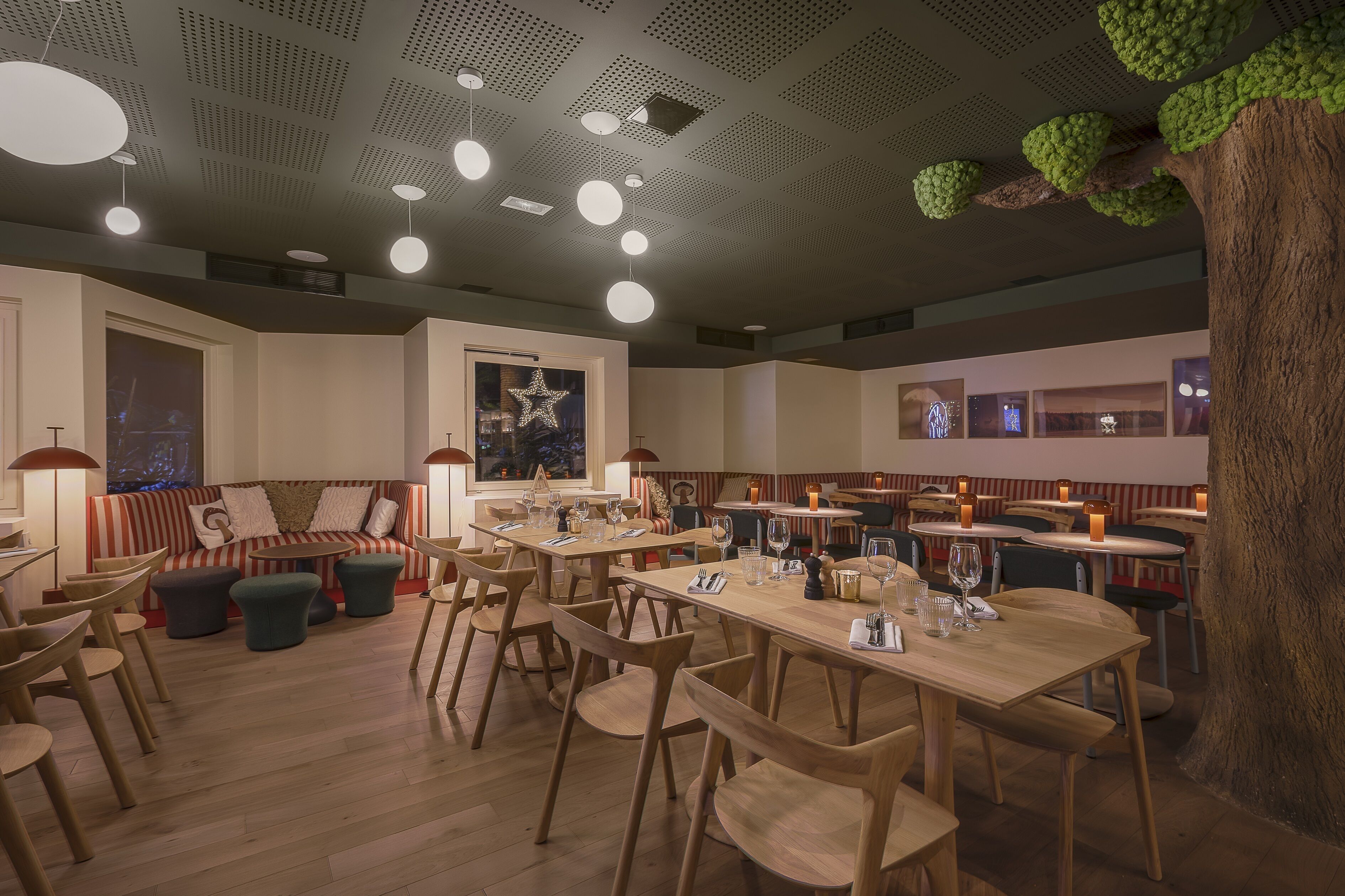 A restaurant with wooden tables and chairs, a large tree trunk, and hanging lights.