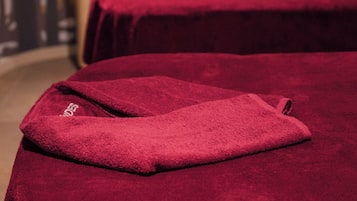 Une chambre avec deux lits recouverts de couvertures rouges, une table de chevet avec une lampe et, au fond, un rideau à rayures.