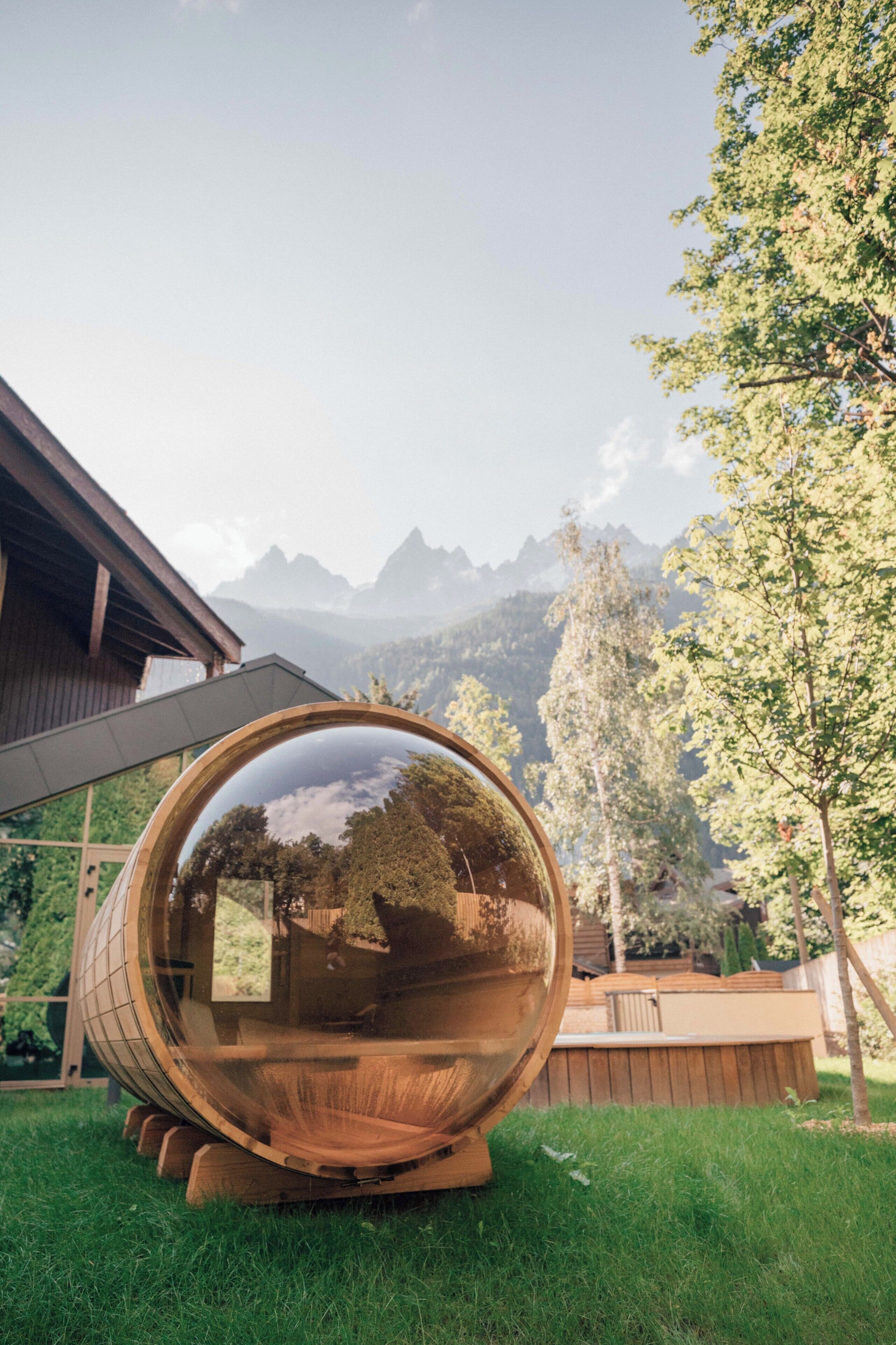 Une grande sphère transparente qui reflète un paysage montagneux, placée sur une pelouse, avec une structure en bois en arrière-plan.
