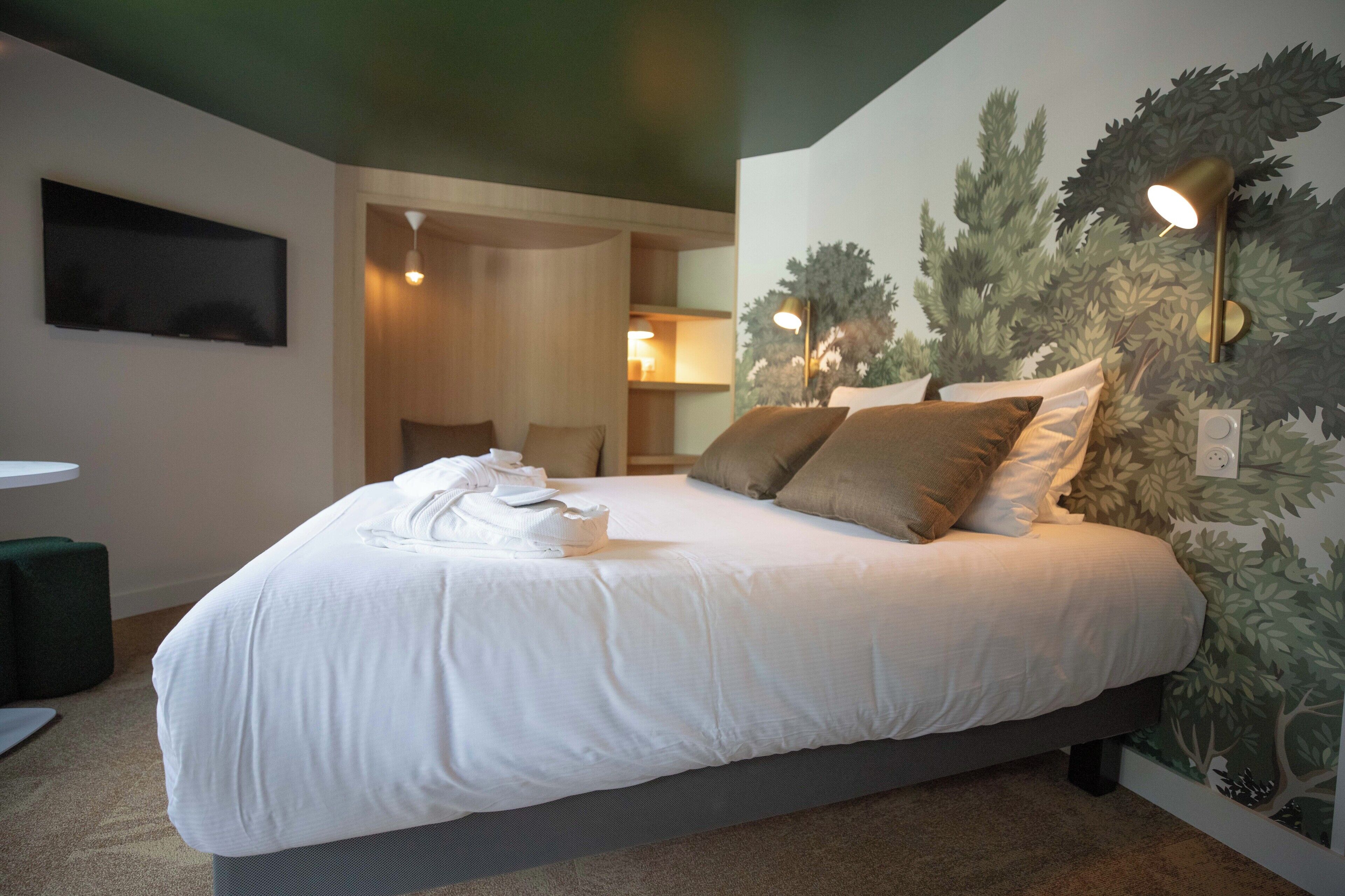 A neatly made bed with pillows, a flat-screen TV on the wall, and a wall-mounted lamp.