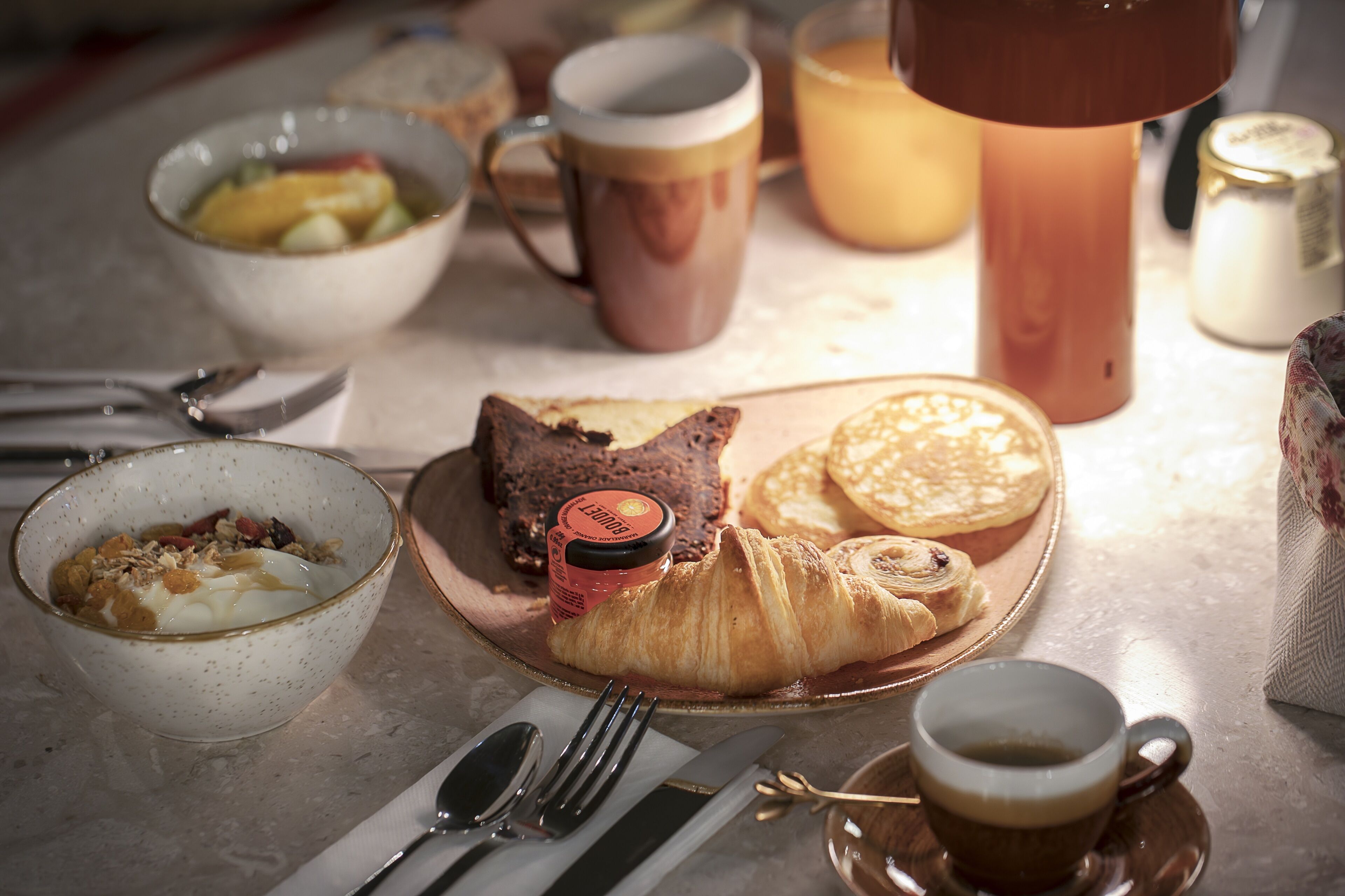 A breakfast setting with a croissant, a bowl of cereal, a mug, and a cup of coffee on a table.