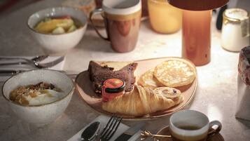 A breakfast setting with a croissant, a bowl of cereal, a mug, and a cup of coffee on a table.