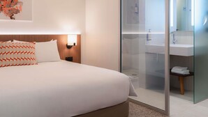 Une chambre d’hôtel moderne avec un grand lit, une table de chevet, une lampe fixée au mur et une porte en verre donnant sur une salle de bain équipée d’un lavabo et d’un miroir.