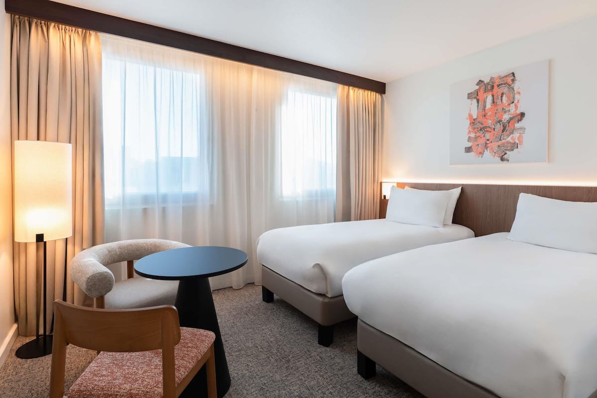 Hotellihuone, jossa on kaksi sänkyä, tuoli, pieni pöytä ja lamppu.