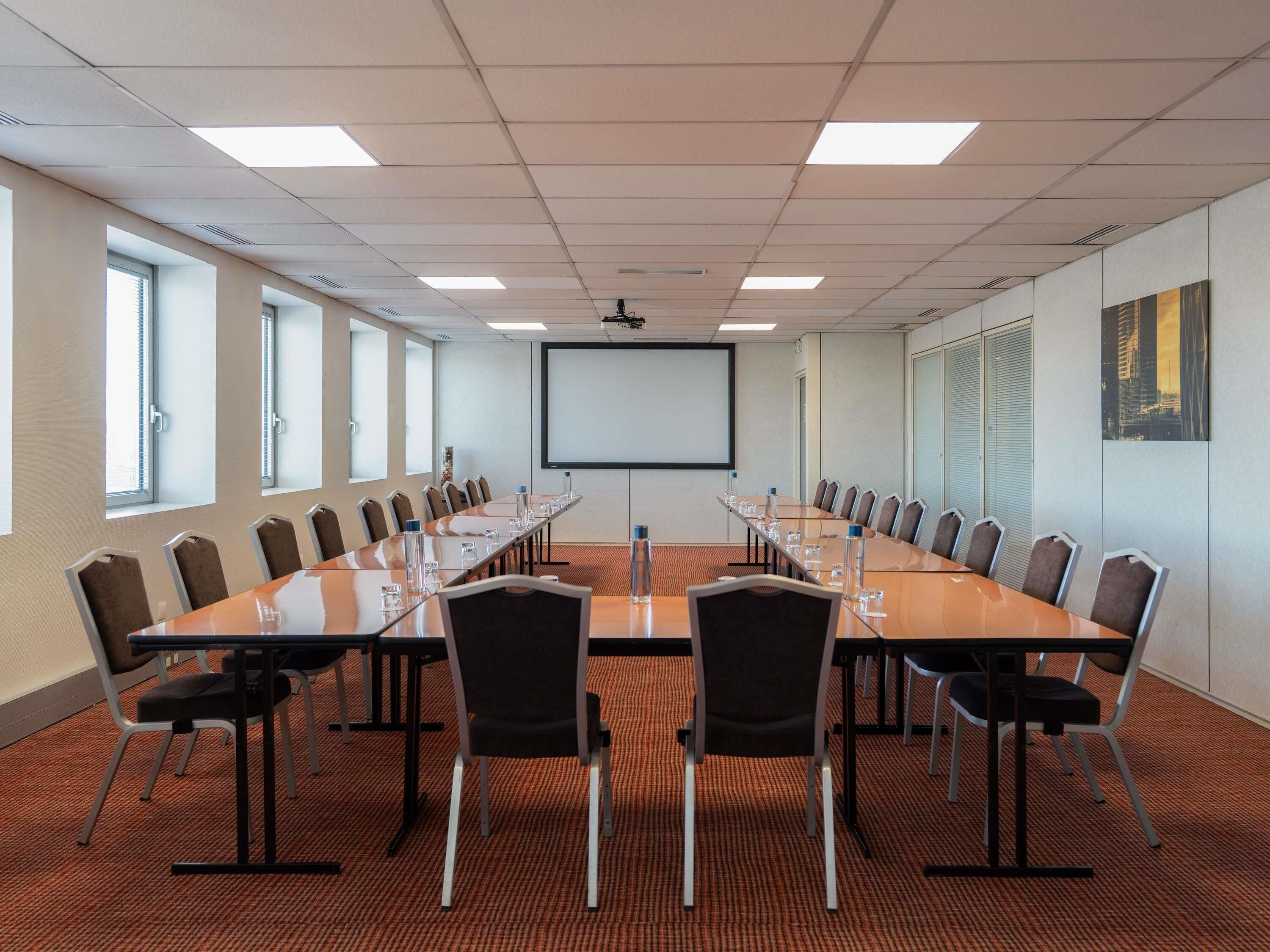 A conference room with a long table, chairs, a projector screen, and windows.