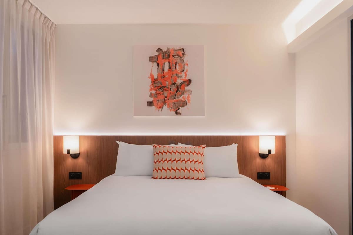 Hotellihuone, jossa on suuri sänky, kaksi yöpöytää, seinälle ripustettu taideteos ja ikkuna, jossa on verhot.