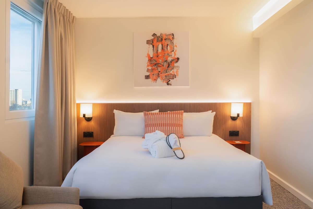Hotellihuone, jossa on suuri sänky, kaksi yöpöytää, sängynpääty, ikkuna verhoilla ja näkymä kaupunkimaisemaan.