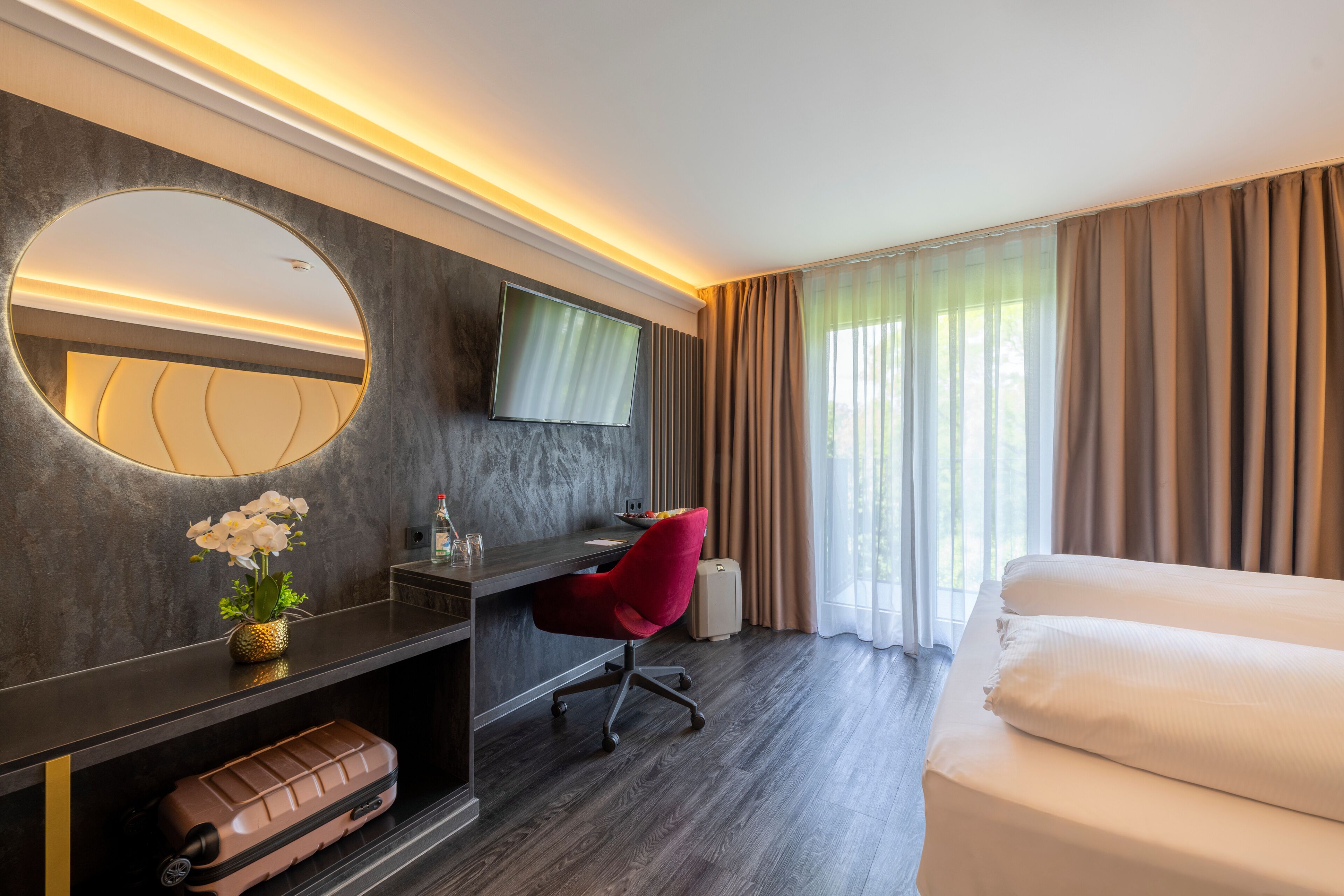 Ein Hotelzimmer mit Bett, Schreibtisch mit Stuhl, Fernseher, Spiegel und einem fenster mit Vorhängen.
