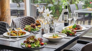 Une table à manger dressée avec des assiettes remplies de nourriture, des verres et une bouteille de vin, entourée de chaises et offrant une vue sur la verdure.