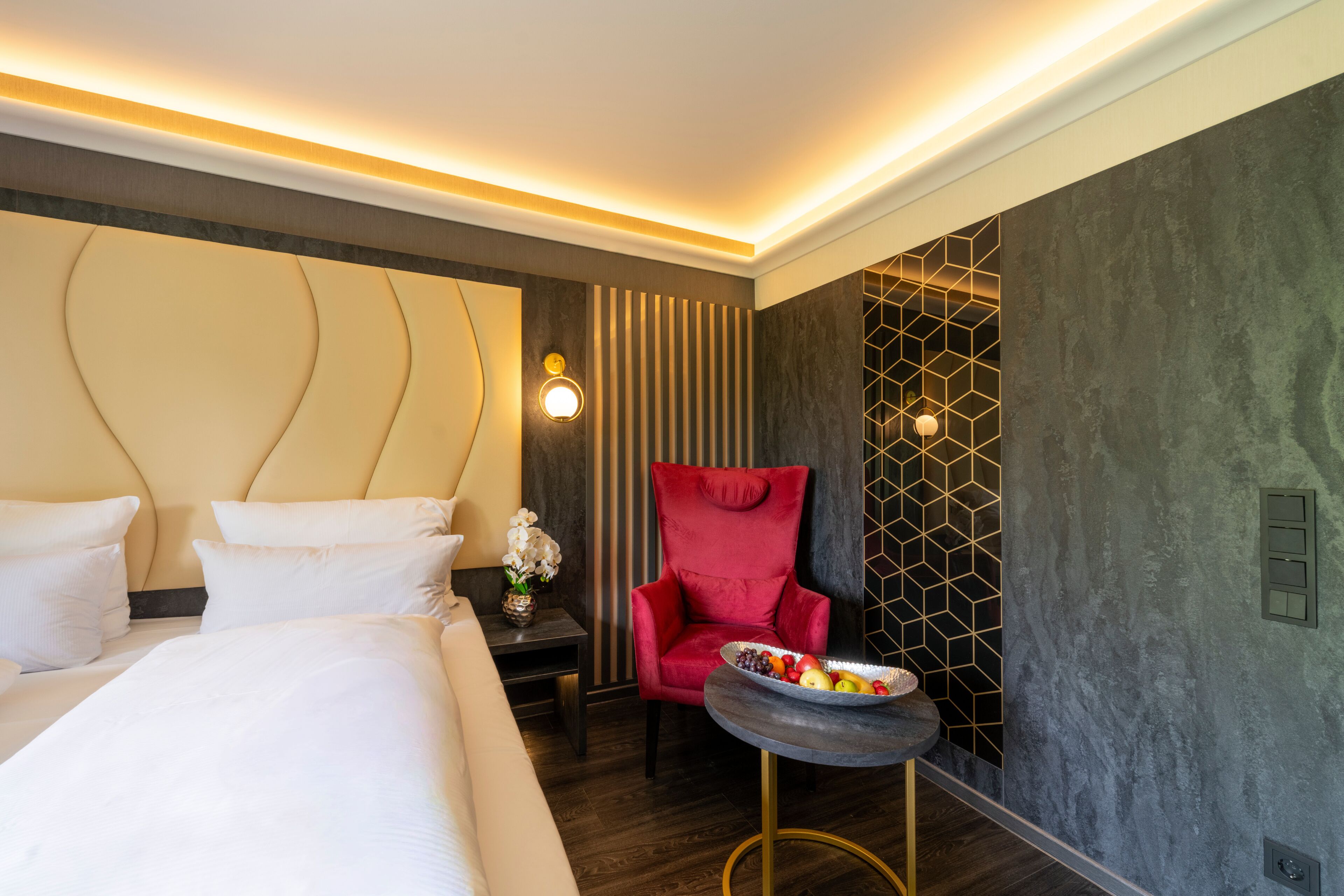 A hotel room with a large bed, a red armchair, a small table with fruit, and a decorative wall panel.