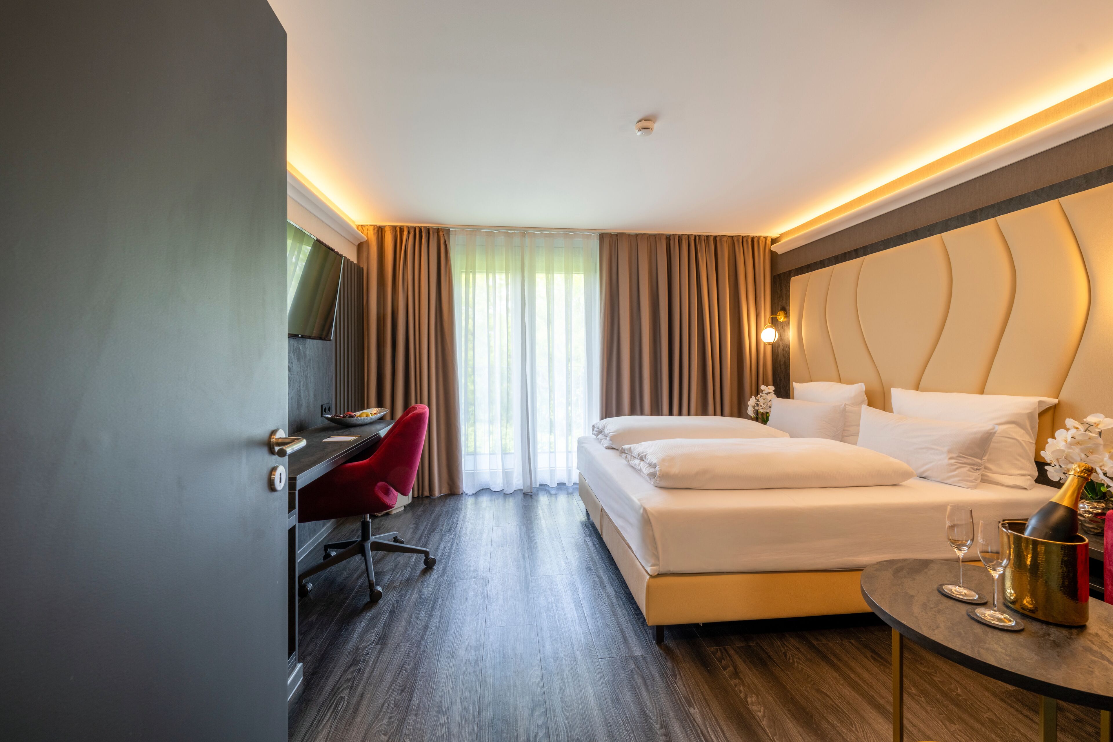 A hotel room with a bed, a desk, a chair, a small table with a bottle and glasses, and a window with curtains.