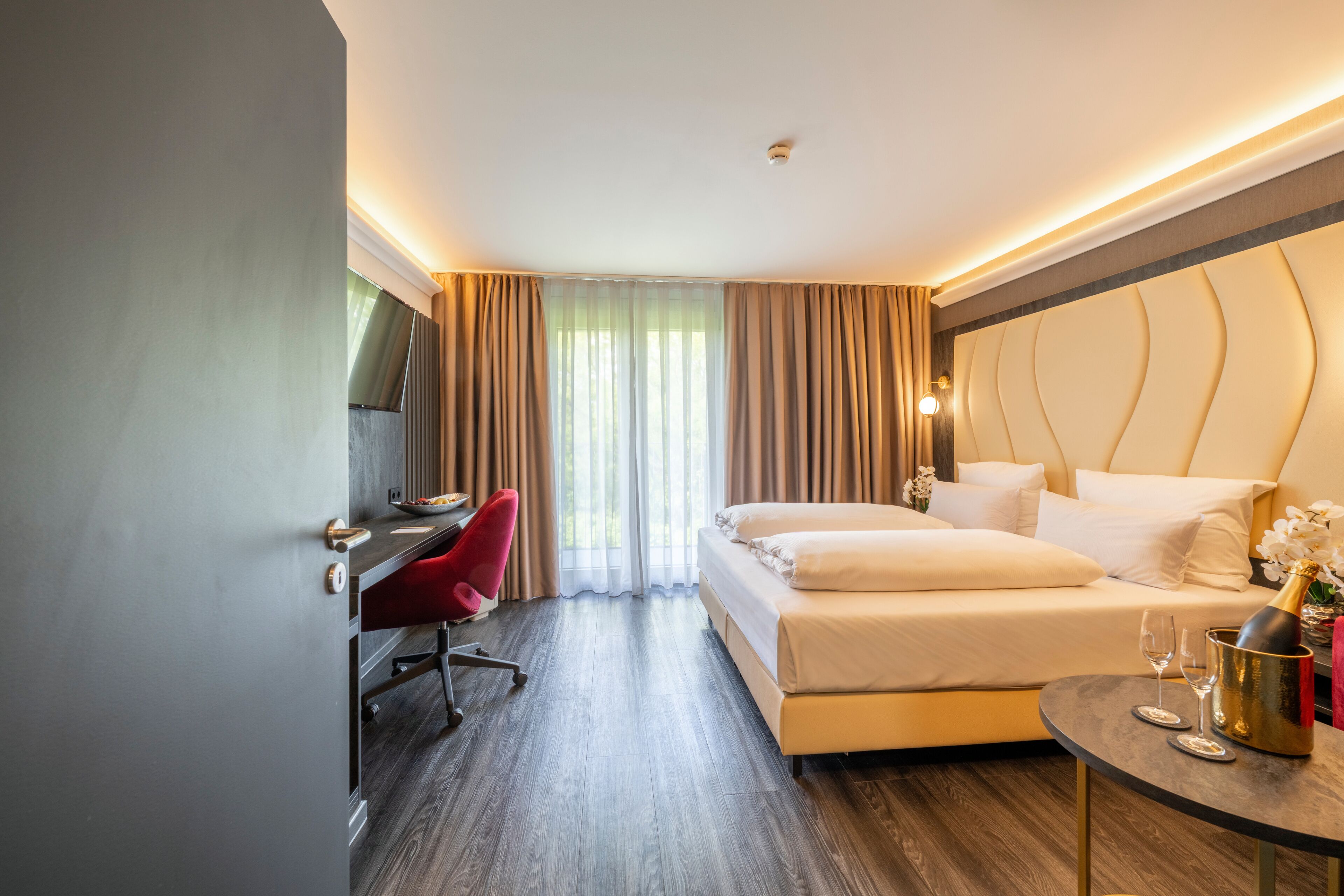 A hotel room with a bed, a desk, a chair, a TV, and a small table with a bottle and glasses.
