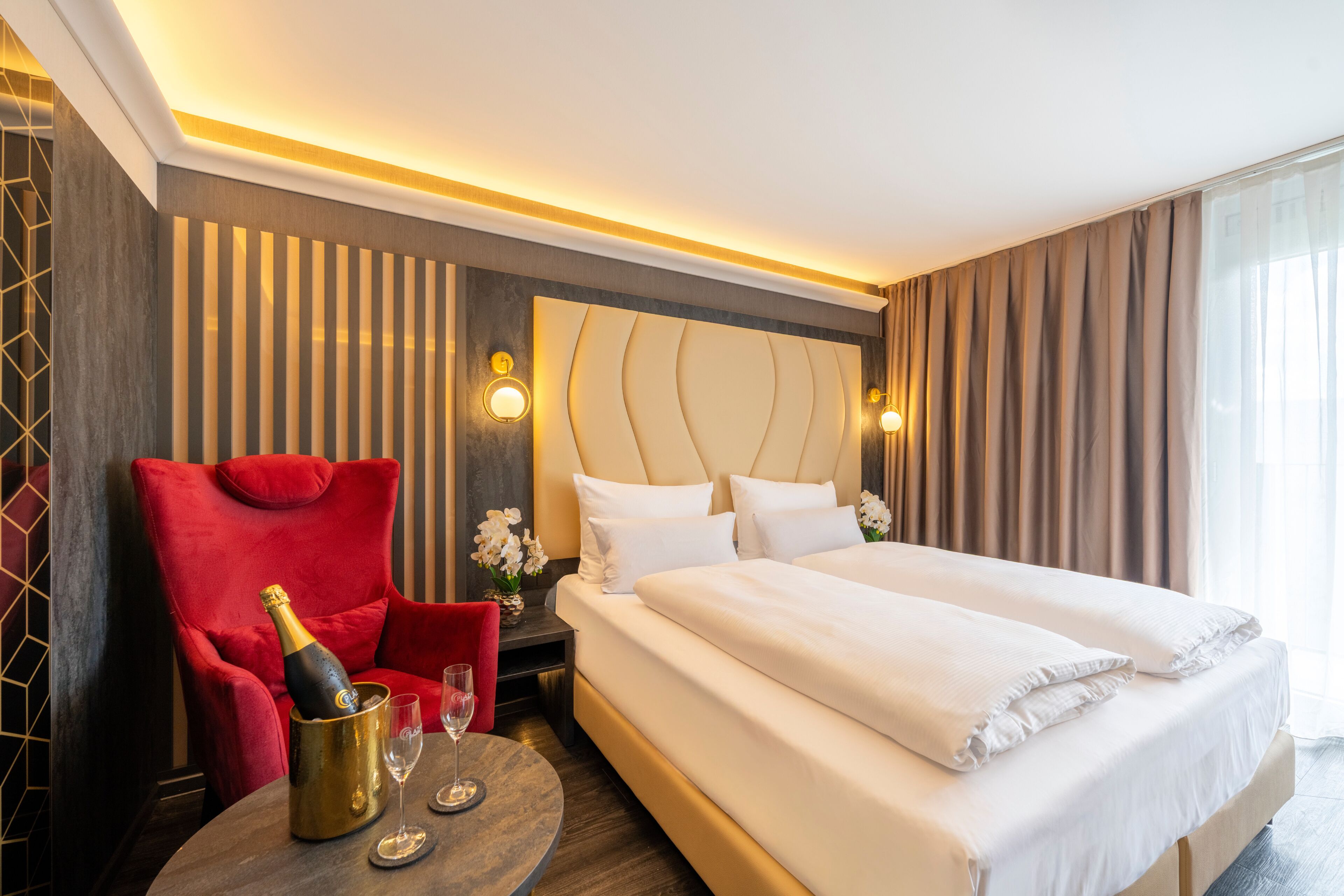 Habitación de hotel con una cama grande, un sillón rojo, una mesita con una botella y vasos, y una mesita de noche con un jarrón de flores.