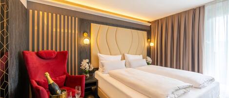 Habitación de hotel con una cama grande, un sillón rojo, una mesita con una botella y vasos, y una mesita de noche con un jarrón de flores.