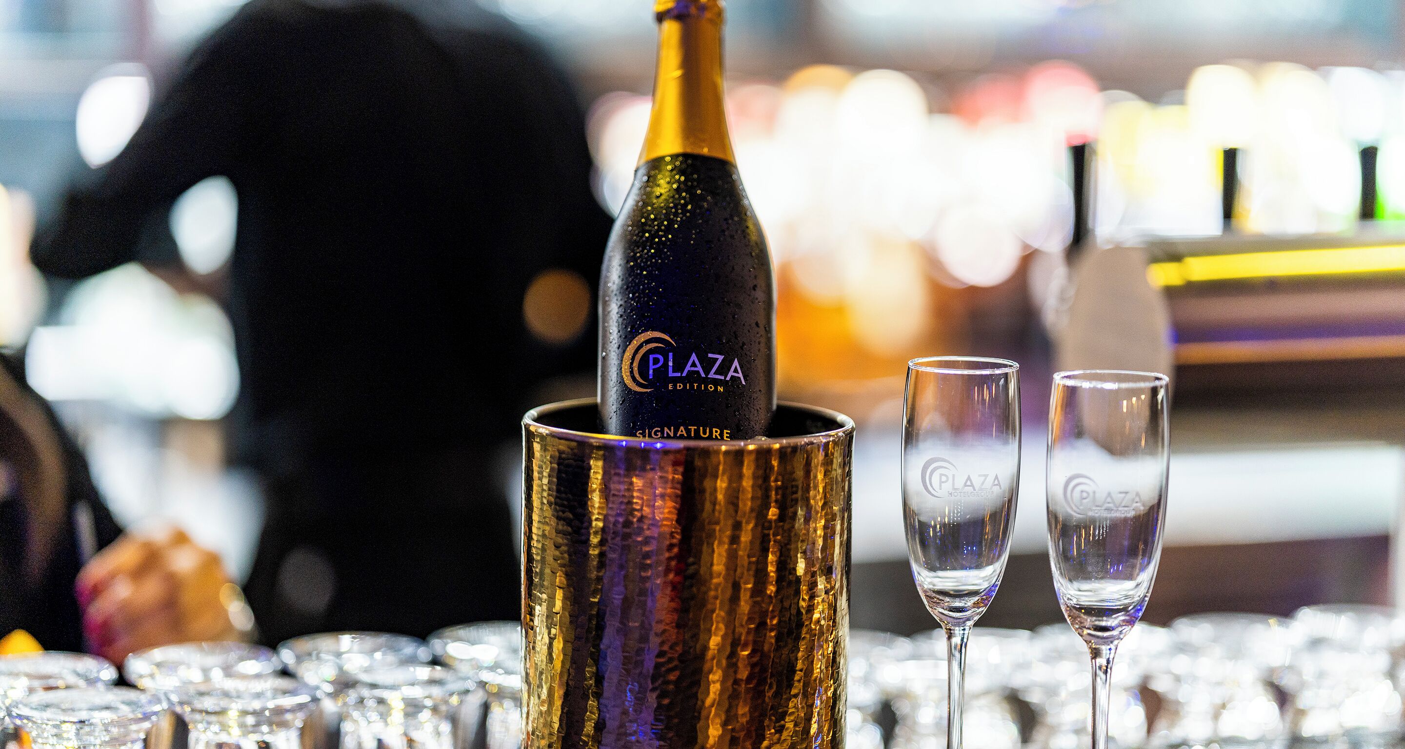A bottle of Plaza Signature Champagne in a golden foil holder with two champagne flutes beside it.