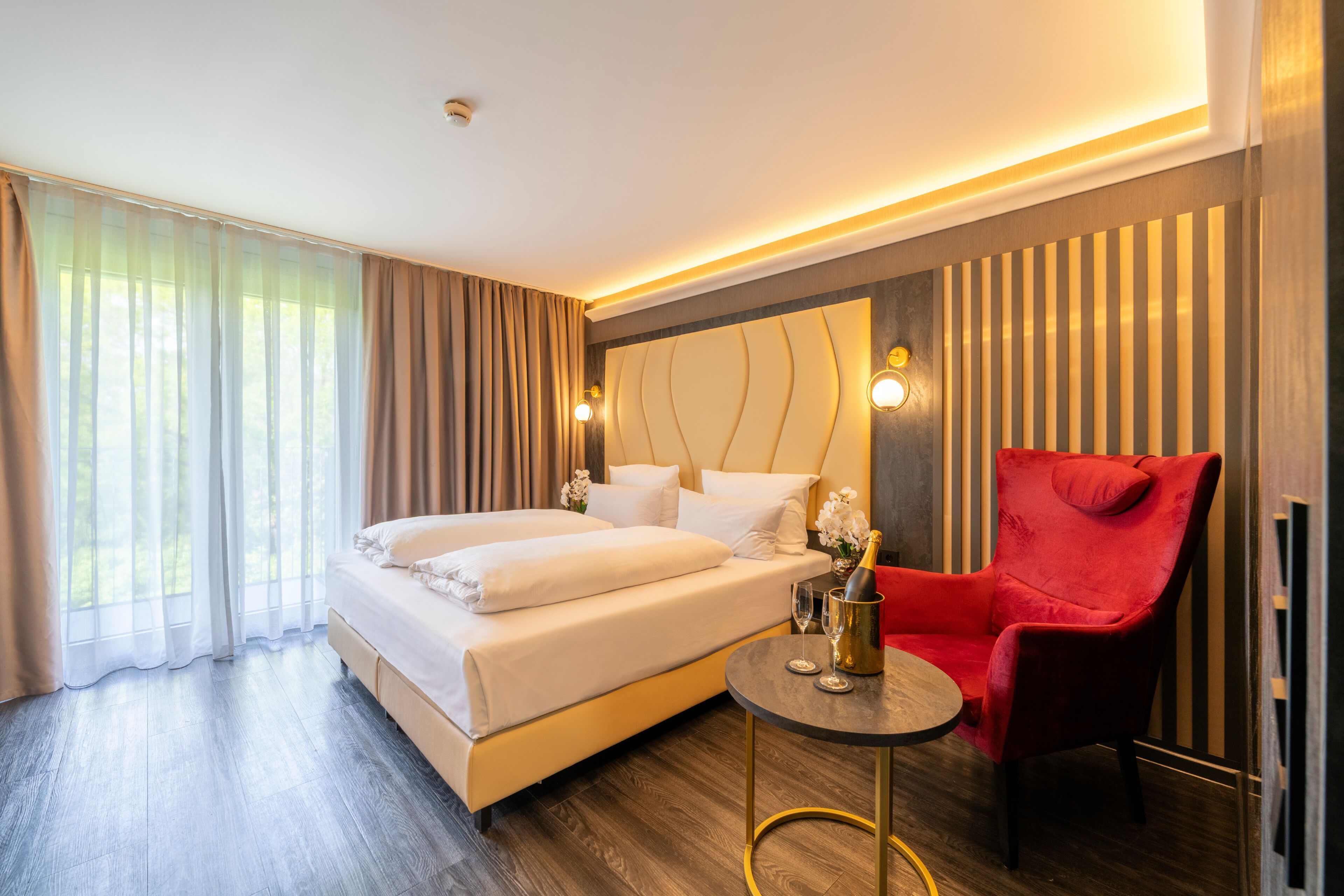 A hotel room with a bed, a red armchair, a small table with a bottle and glass, and a large window with curtains.