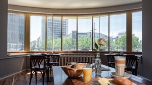 A dining area with a table set for breakfast, including bread, fruit, and beverages, with a view of buildings and trees through large windows. - acom Hotel Berlin Kurfürstendamm (Berlin)