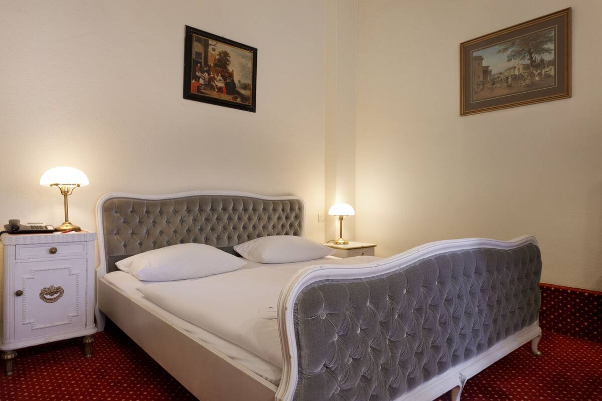 Una cama bien hecha con un cabecero capitonado, una mesita de noche con lámpara y dos cuadros enmarcados en la pared.