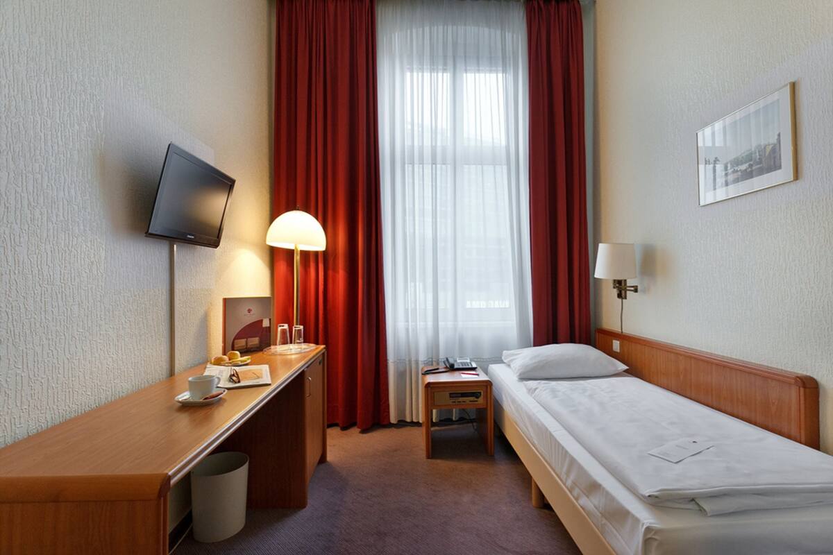 Habitación de hotel con cama, escritorio con lámpara, televisión y ventana con cortinas.