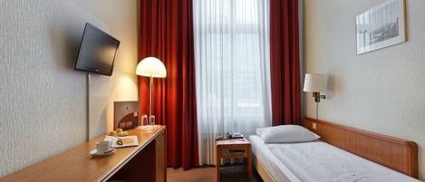 A hotel room with a bed, a desk with a lamp, a TV, and a window with curtains.