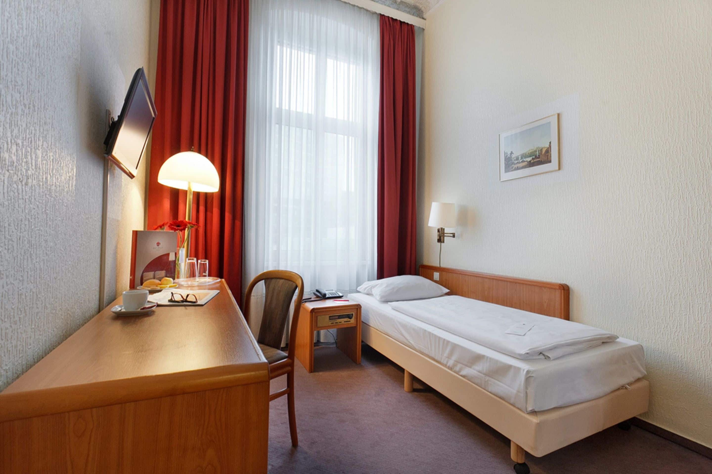 Ein Hotelzimmer mit Holzschreibtisch, einem Sessel, einem Bett, einem Nachttisch, einer Lampe, einem Fernseher und einem Fenster mit roten Vorhängen.