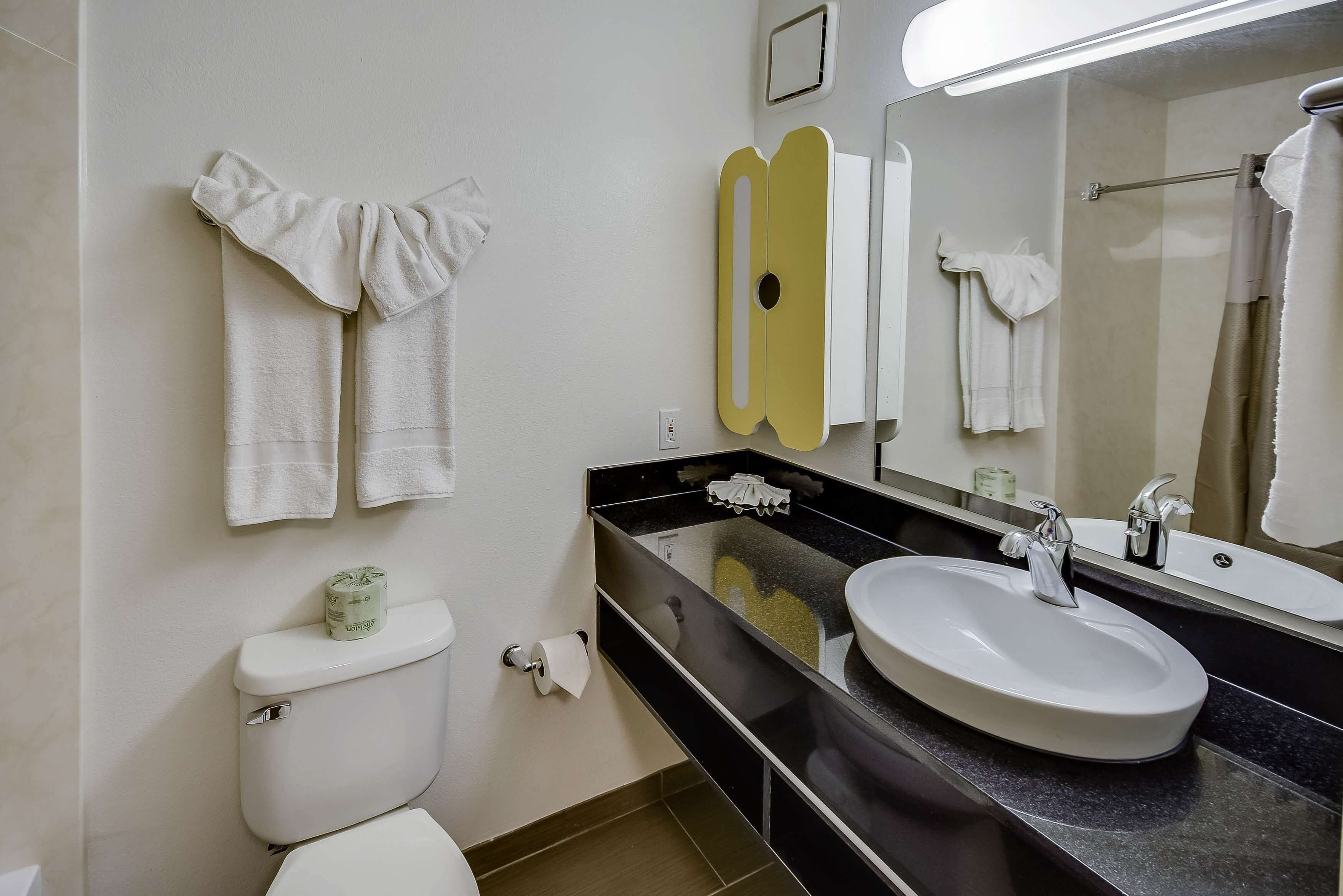 A bathroom with a toilet, sink, and mirror. There are towels hanging on the wall and a roll of toilet paper on the counter.