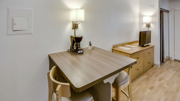 A small dining area with a wooden table, two chairs, a coffee maker, a television on a wooden cabinet, and wall-mounted lights.