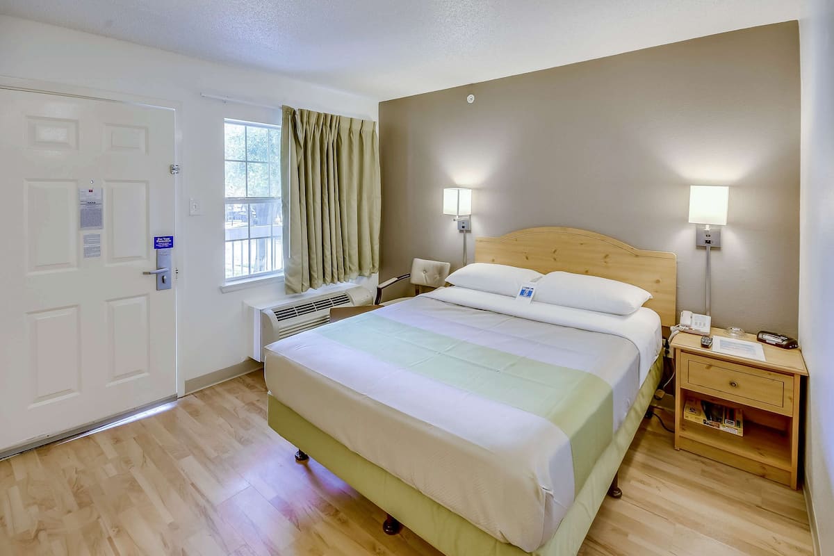 A hotel room with a bed, bedside lamps, a wooden nightstand, a window with curtains, and a door.