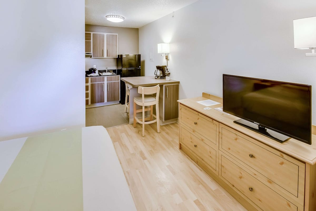A compact hotel room with a bed, a TV, a desk with a chair, and a kitchenette in the background.