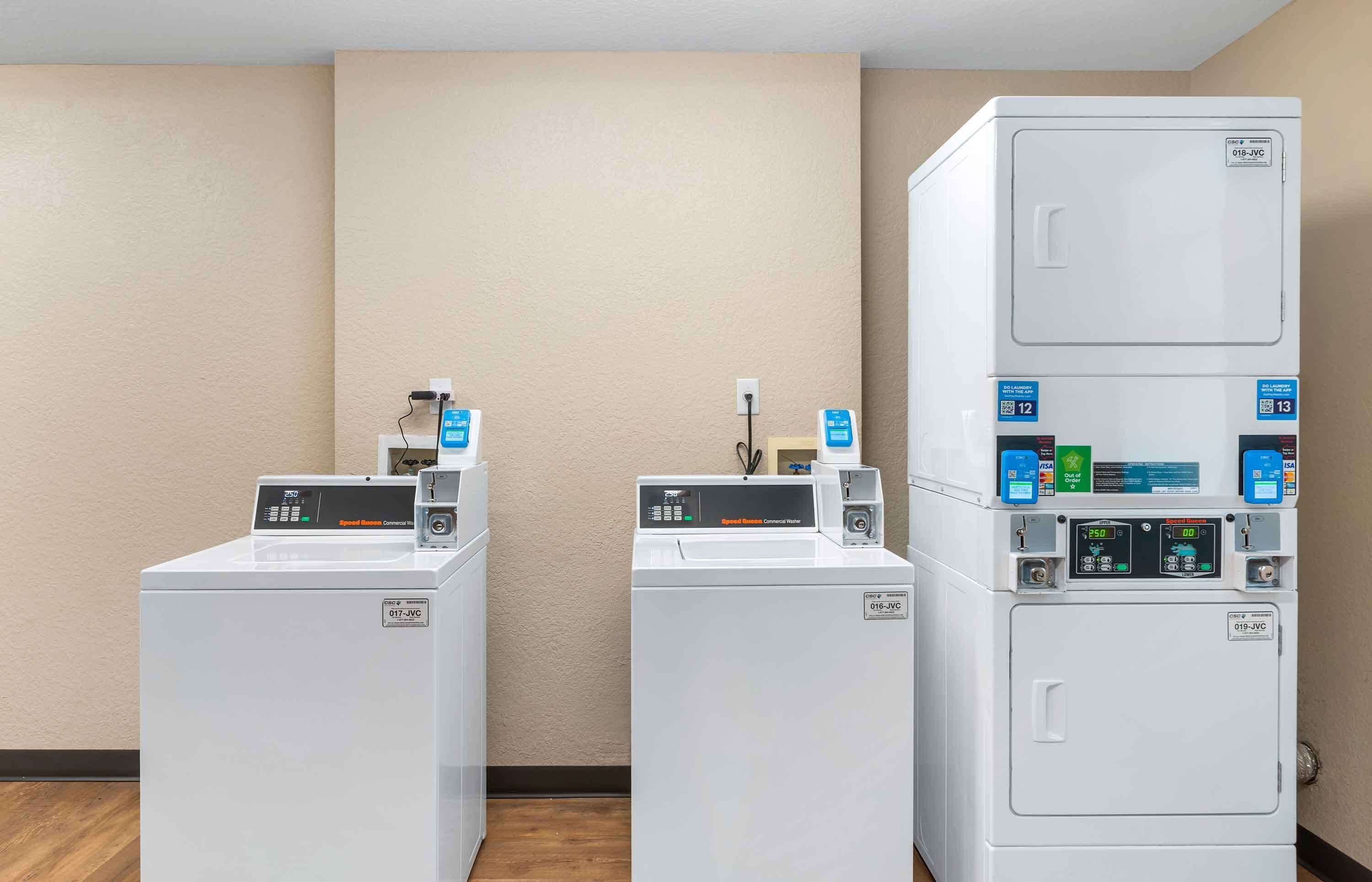 Three front-loading washing machines in a hotel room, each with a control panel and a small shelf above.