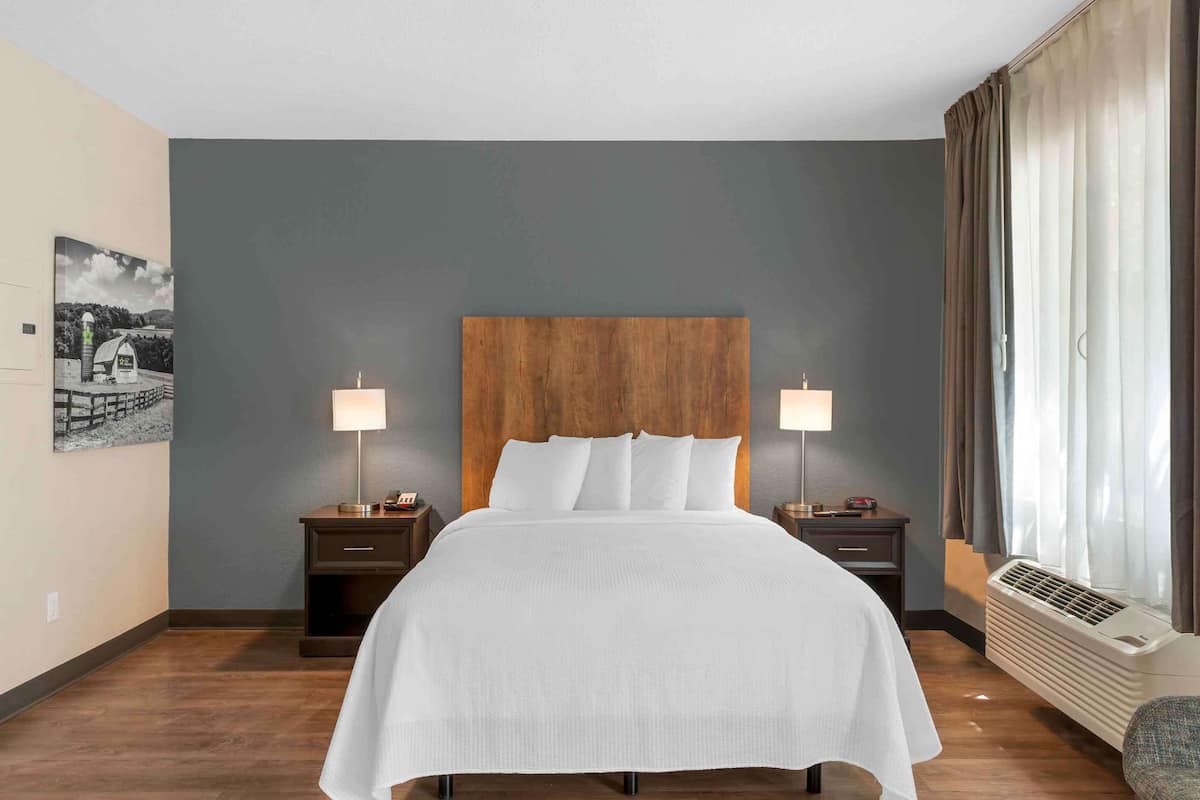 Ein Hotelzimmer mit einem großen Bett, zwei Nachttischen mit Lampen, einer Wand-Klimaanlage und einem Fenster mit Vorhängen.