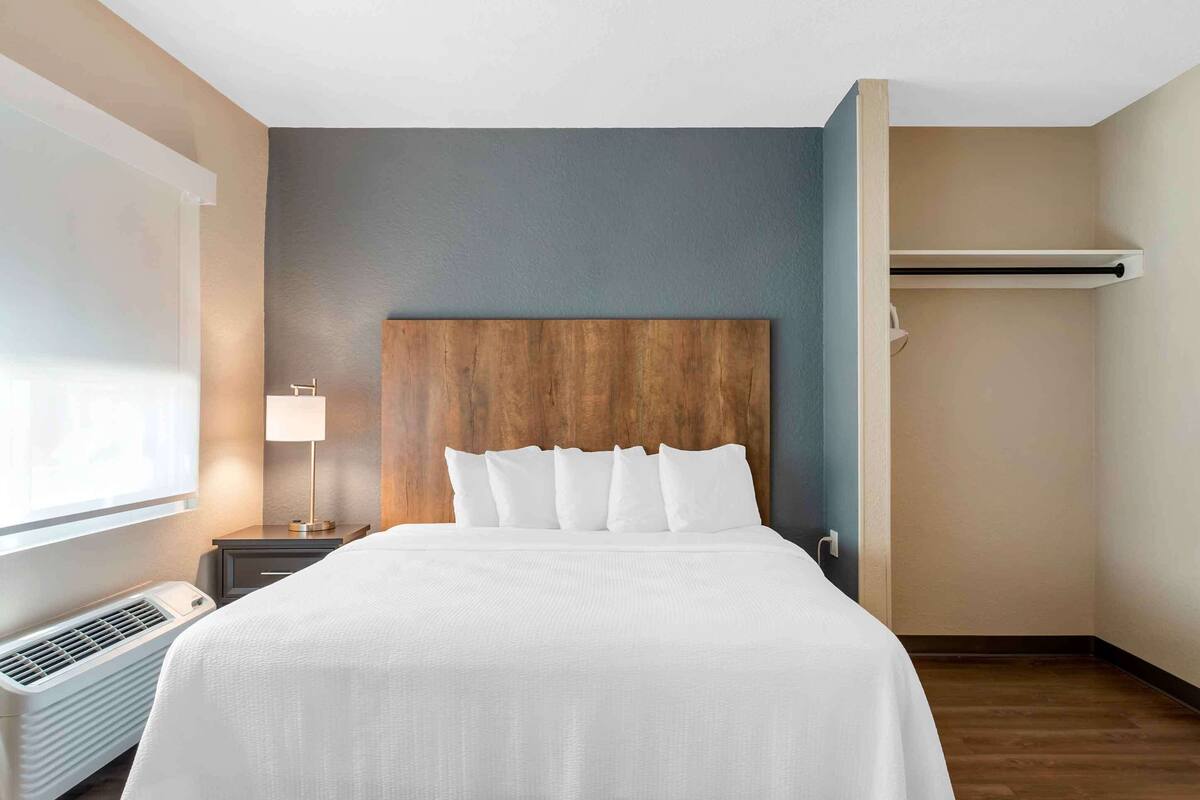 Ein Hotelzimmer mit einem großen Bett, einem Nachttisch mit Lampe, einer Wand-Klimaanlage und einem Fenster mit Jalousien.