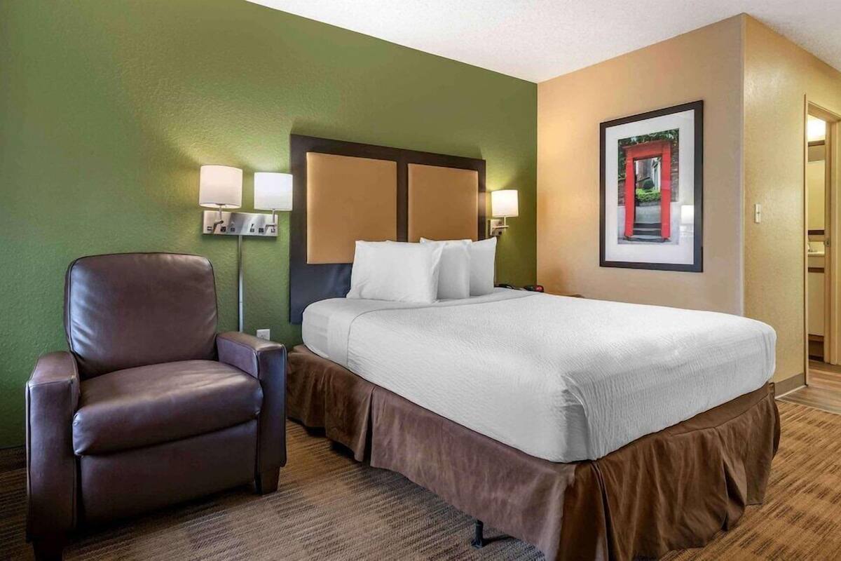 A hotel room with a bed, a chair, a nightstand, and a wall-mounted lamp.
