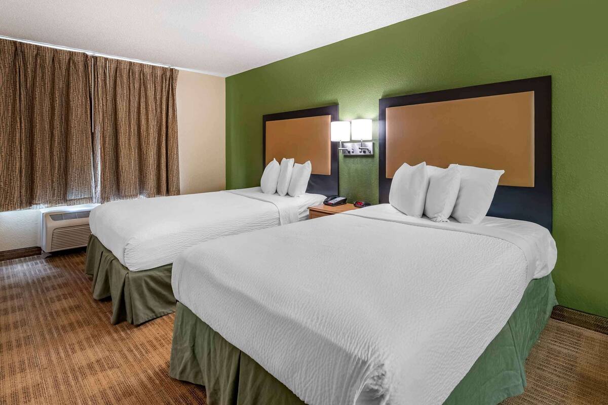 A hotel room with two beds, a green wall, a window with curtains, and a bedside lamp.