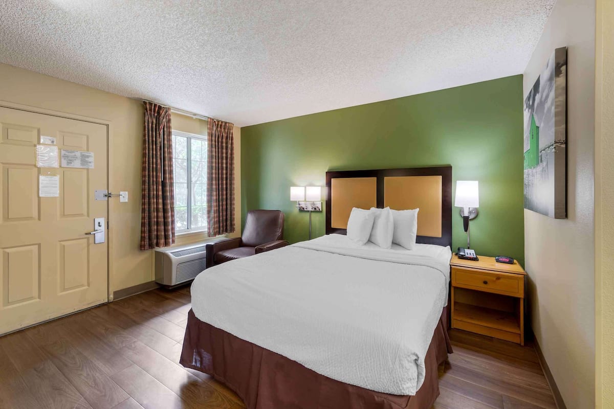 A hotel room with a large bed, a chair, a nightstand, a lamp, and a painting on the wall.