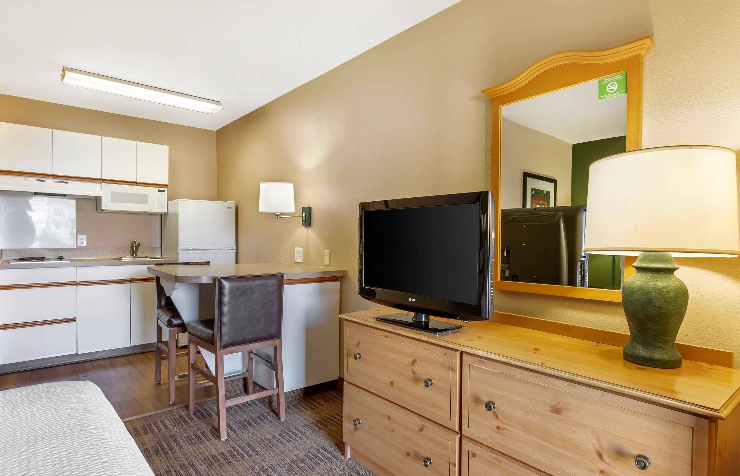 A hotel room with a kitchenette, a flat-screen TV, a mirror, a lamp, and a bed.