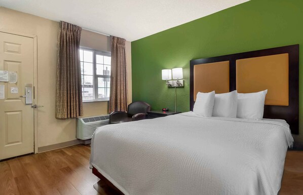 A hotel room with a large bed, a desk with a chair, a window with curtains, and a green wall.