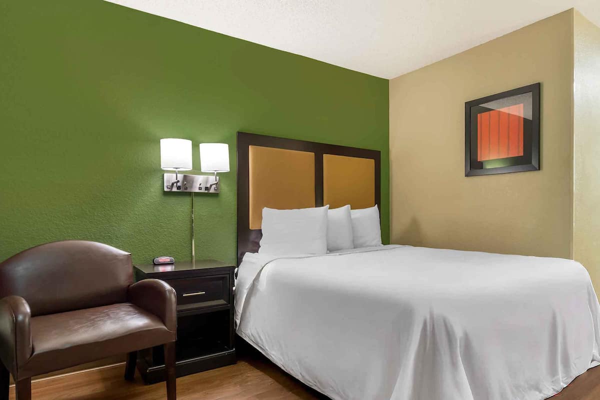 Une chambre d’hôtel avec un lit, une chaise, une table de chevet et une lampe fixée au mur.