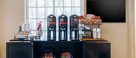 Una estación de café con varios dispensadores, una selección de pasteles y una pizarra con el menú.