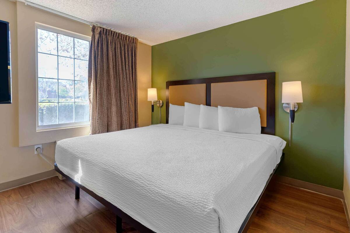 A hotel room with a large bed, two bedside lamps, a window with curtains, and a green wall.