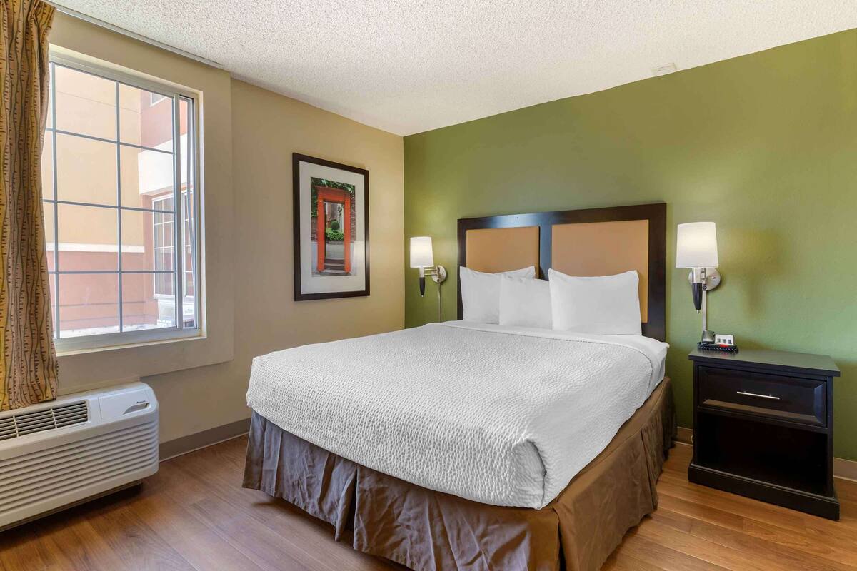 A hotel room with a large bed, two bedside lamps, a nightstand, a window with curtains, a wall-mounted air conditioner, and a framed picture on the wall.