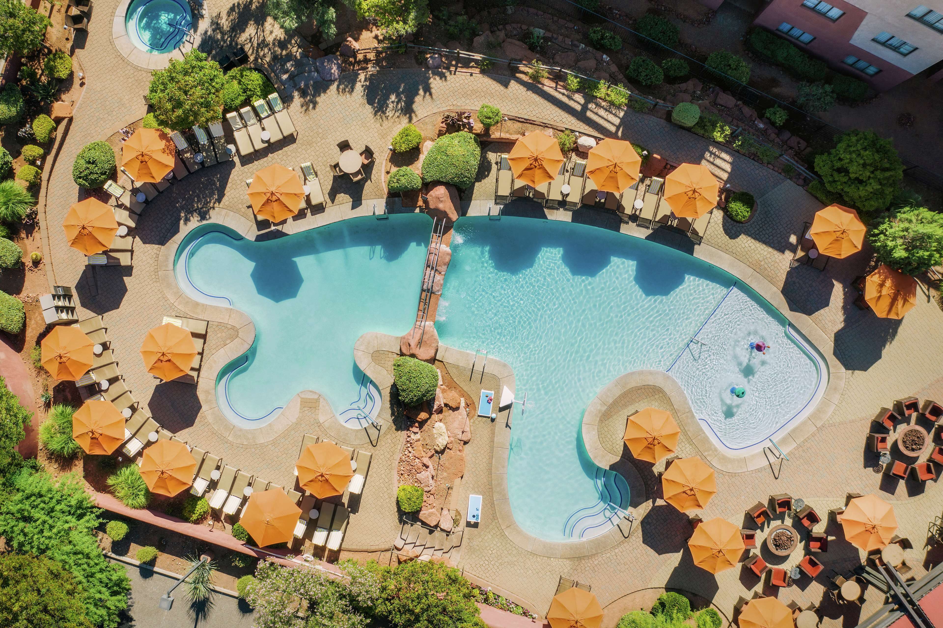 An aerial view of a resort with a large, uniquely shaped swimming pool surrounded by sun umbrellas and lounge chairs.