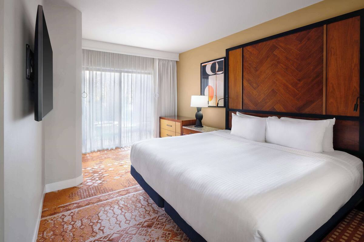 A hotel room with a large bed, a flat-screen TV mounted on the wall, a nightstand with a lamp, a wooden headboard, and a large window with sheer curtains.