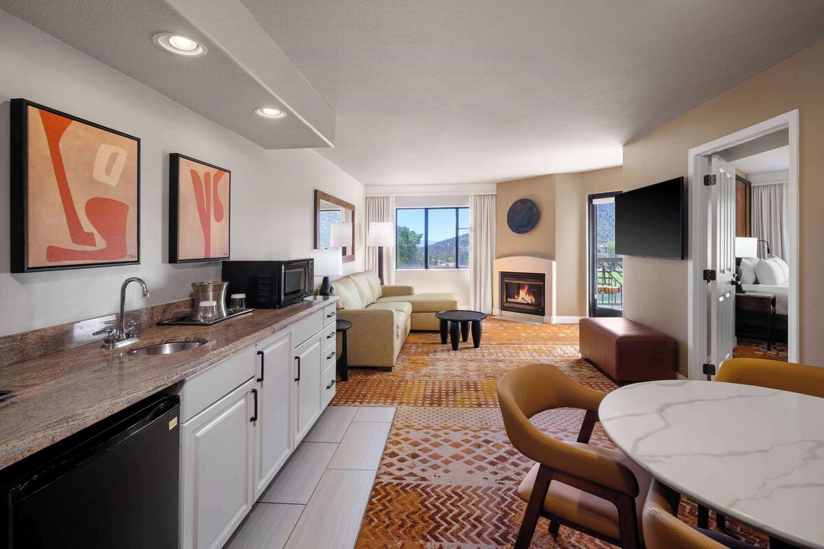 A modern kitchen with a granite countertop, stainless steel sink, and a built-in dishwasher. There is a seating area with a sofa and a coffee table, and a fireplace in the background.
