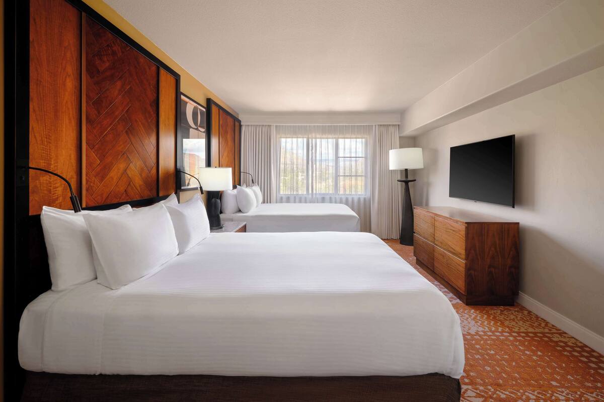 A hotel room with two beds, a TV, a dresser, and a window with curtains.