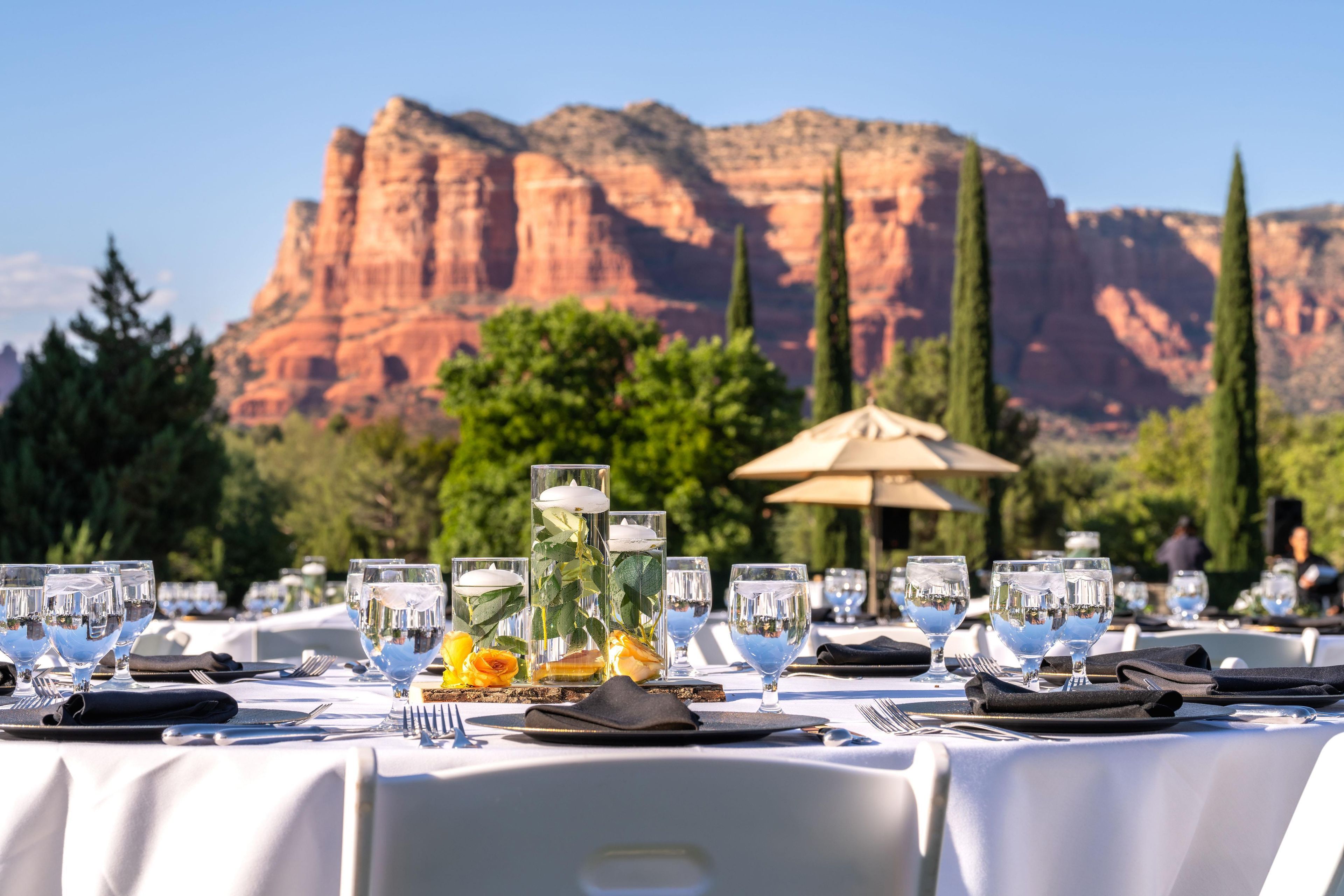 An outdoor dining area with tables set for a meal, featuring clear glassware and a decorative centerpiece.