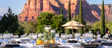 An outdoor dining area with tables set for a meal, featuring clear glassware and a decorative centerpiece.