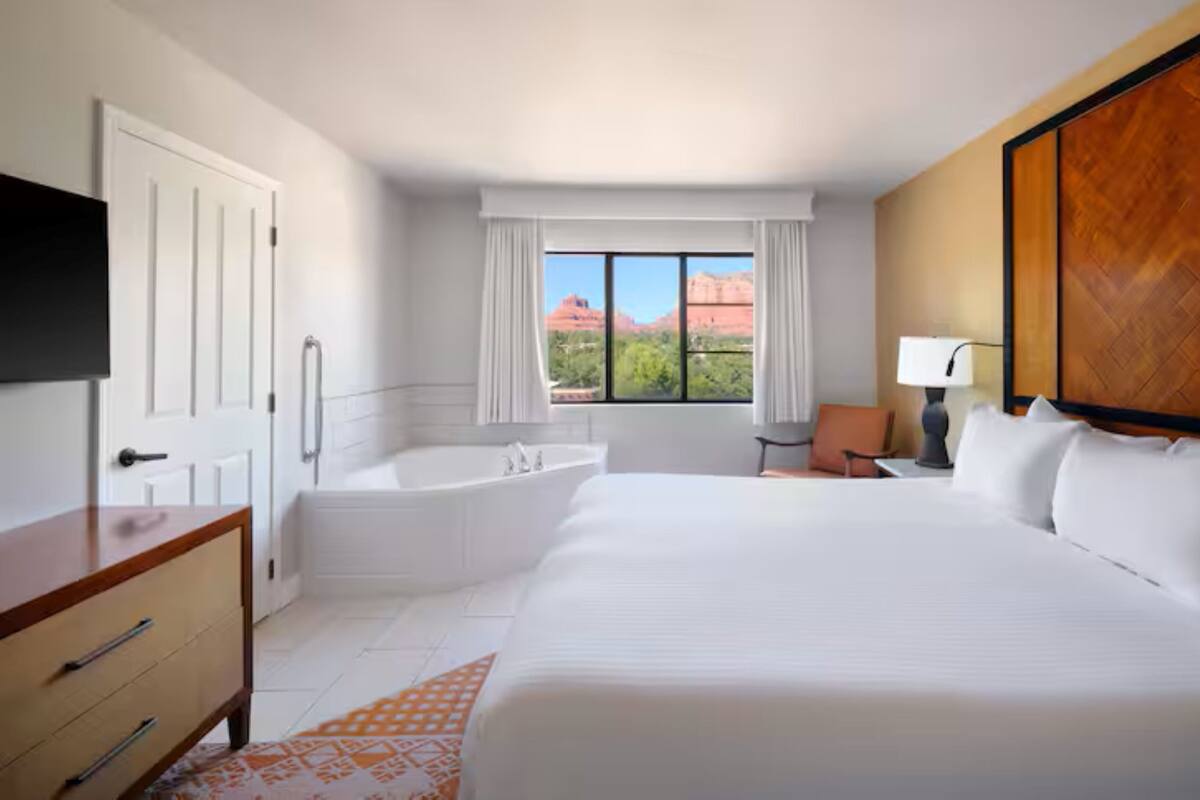 A hotel room with a large bed, a separate bathroom with a bathtub, a desk, a chair, a TV mounted on the wall, and a window with a view of red rock formations.