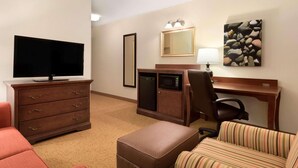 A hotel room with a flat-screen TV, a wooden desk with a lamp, a striped chair, a brown ottoman, and a large mirror.