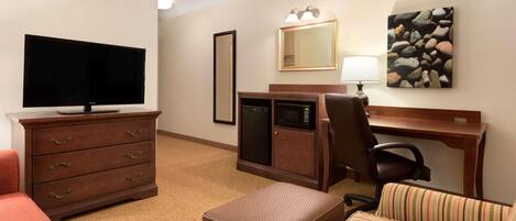 A hotel room with a flat-screen TV, a wooden desk with a lamp, a striped chair, a brown ottoman, and a large mirror.